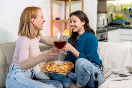 Two best friends are enjoying a fun and cozy night in together, sitting in the living room with pizza and red wine.の写真素材