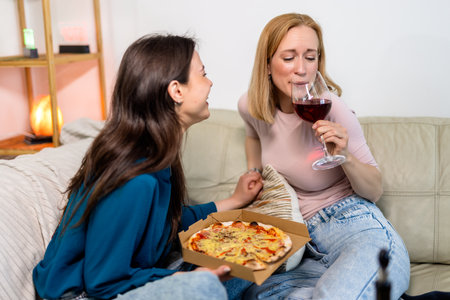Two best friends are enjoying a fun and cozy night in together, sitting in the living room with pizza and red wine.の写真素材