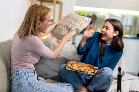 Two best friends are having the ultimate girls' night in, complete with pizza, red wine, and an unexpected pillow fight in the living room. Bursting into laughter and silliness, they enjoy every carefree moment together, surrounded by comfort and joy.の写真素材