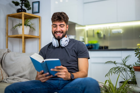 A young bearded man sits comfortably on a sofa in a cozy living room, fully absorbed in a book. Surrounded by the quiet of home, he enjoys a peaceful moment of solitude, trading screen time for pages.の写真素材