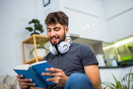 A young bearded man sits comfortably on a sofa in a cozy living room, fully absorbed in a book. Surrounded by the quiet of home, he enjoys a peaceful moment of solitude.の写真素材
