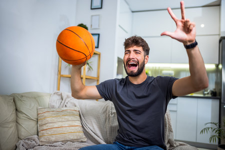 A young man is sitting in his living room, fully engrossed in a basketball game. With a basketball in his hand.の写真素材
