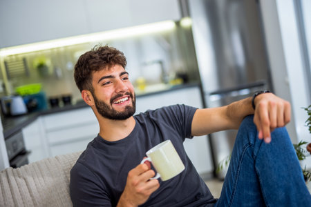 A young bearded man sits comfortably on a sofa in his cozy living room, enjoying a warm cup of coffee. It's a simple but peaceful moment of relaxation, with the quiet atmosphere of home providing the perfect setting for a slow, reflective break.の写真素材