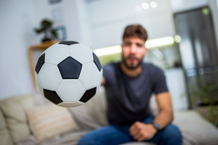 A young man sits in his living room, fully immersed in a soccer match, cheering with excitement as his favorite team plays. Holding a soccer ball in his hand, he expresses his passion for the sport and team spirit.の写真素材