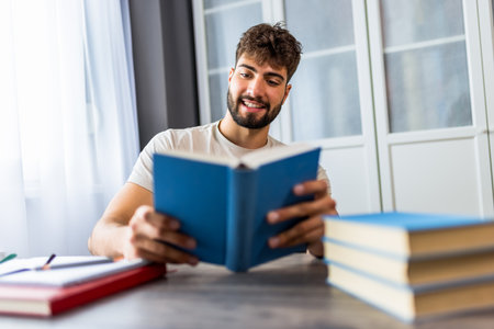 A young student sits at a desk surrounded by books, fully immersed in studying for an upcoming exam. His focused expression shows the dedication required as he works through his textbooks and notes.の写真素材