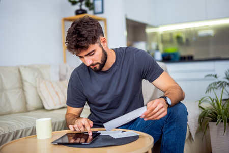 A young bearded man sits in his living room, surrounded by a pile of bills. With a worried expression, he struggles to keep up with the financial pressures of everyday life.の写真素材