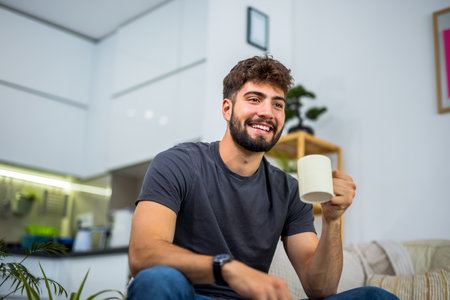 A young bearded man sits comfortably on a sofa in his cozy living room, enjoying a warm cup of coffee. It's a simple but peaceful moment of relaxation, with the quiet atmosphere of home providing the perfect setting for a slow, reflective break.の写真素材
