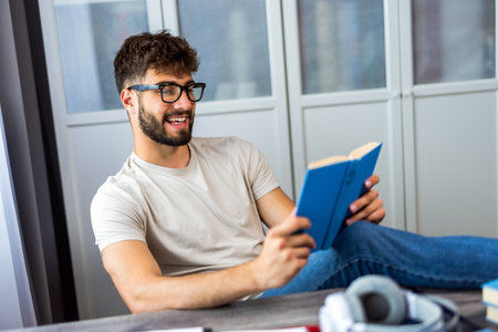 Young student sits comfortably with a book in hand, wearing glasses as he immerses himself in reading. His deep concentration and thoughtful expression highlight the joy of learning and expanding knowledge.の写真素材
