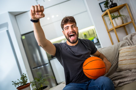 A young man is sitting in his living room, fully engrossed in a basketball game. With a basketball in his hand.の写真素材