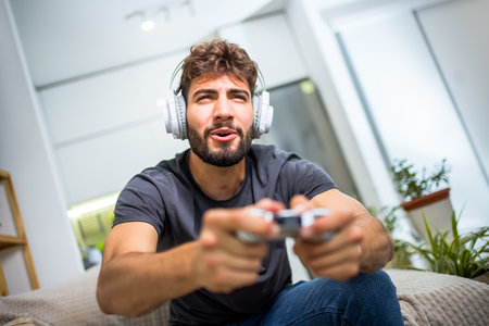 A young man sits comfortably at home, fully immersed in his gaming experience, holding a controller and focused on the screen in front of him. With his favorite game in play, heâs fully engaged in the digital world, living out virtual adventures and challenges.の写真素材