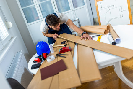 A young man works diligently on renovating his apartment, focusing on the installation of laminate flooring. With the right tools in hand, he carefully measures and fits the laminate panels into place, transforming the space with his DIY skills.の写真素材