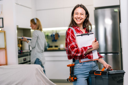 A woman in the foreground holds a clipboard and tools, looking focused and ready to work. Behind her, another woman is using a caulking gun to apply silicone. The image shows teamworkの写真素材