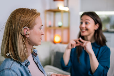 Two young girls are using sign language to talk to each other. One of them is wearing a hearing aid. They are smiling and enjoying the conversation.の写真素材