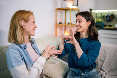 Two young girls are using sign language to talk to each other. One of them is wearing a hearing aid. They are smiling and enjoying the conversation.の写真素材