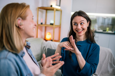 Two young girls are using sign language to talk to each other. One of them is wearing a hearing aid. They are smiling and enjoying the conversation.の写真素材