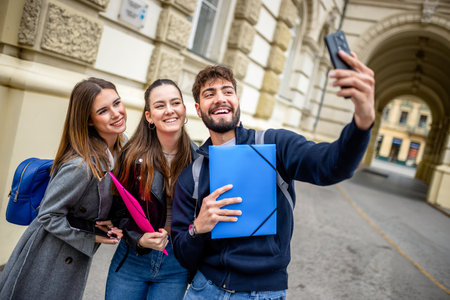 Three cheerful students, holding books and a folder, gather closely together outdoors as they take a selfie using a smartphone.の写真素材