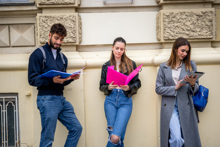 Three students sit outdoors in a casual setting, each focused on their own studies while sharing the same space. They hold books, notebooks, or tablets, emphasizing independent learning within a communal environment.の写真素材