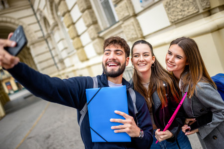 Three cheerful students, holding books and a folder, gather closely together outdoors as they take a selfie using a smartphone.の写真素材