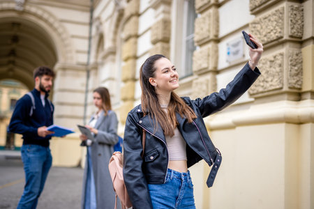 A smiling student holds a stack of books and takes a selfie with a smartphone, capturing a moment of joy and connection.の写真素材
