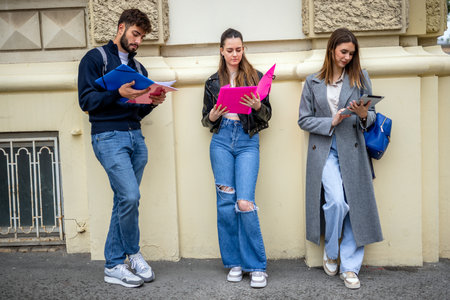 Three students sit outdoors in a casual setting, each focused on their own studies while sharing the same space. They hold books, notebooks, or tablets, emphasizing independent learning within a communal environment.の写真素材