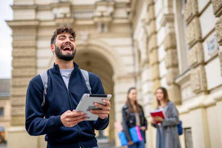 A cheerful student stands in the foreground, smiling confidently at the camera while holding a tablet. Two fellow students are seen slightly blurred in the background, engaged in conversation or study.の写真素材