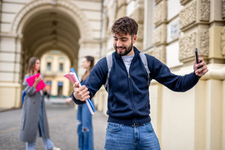 A smiling student holds a stack of books and takes a selfie with a smartphone, capturing a moment of joy and connection. Behind him, two classmates are visible, casually interacting and enjoying their time together.の写真素材