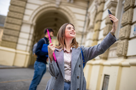A smiling student holds a stack of books and takes a selfie with a smartphone, capturing a moment of joy and connection.の写真素材