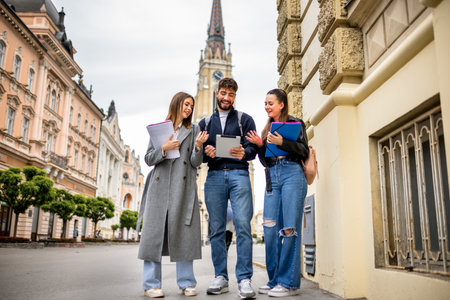 A group of three teenage students walking down a city street, studying together. They are holding books and a tablet, discussing and sharing educational content as they go.の写真素材