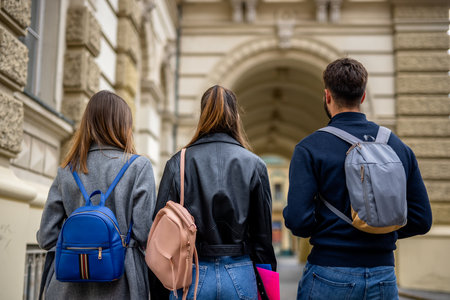 A rear view of three teenage students walking down a city street. They are carrying backpacks and books, dressed casually, and appear to be heading to or from school or a study session.の写真素材