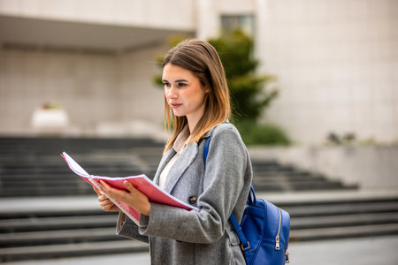 A young female student standing outside and reading a book on a sunny day. She is focused and immersed in her studies.の写真素材