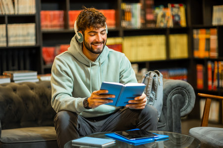 A male student sits in a quiet library, studying from a physical book while wearing headphones,...