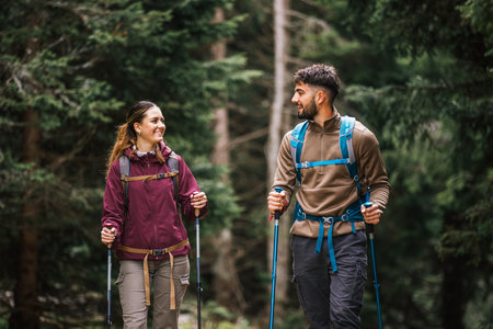 A young man and woman, fully equipped with backpacks, hiking poles, and outdoor clothing, walk...