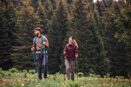 A young man and woman, fully equipped with backpacks, hiking poles, and outdoor clothing, walk...