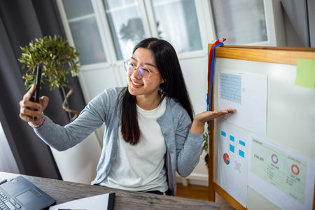 A young Thai woman works from home, delivering an online presentation using her smartphone. She actively engages her audience by pointing to a presentation displayed on a board beside her.の写真素材