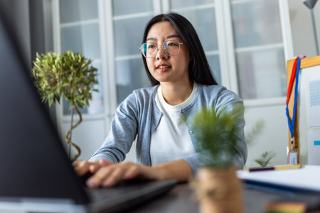 A young Thai woman is working from home, focused on her computer in a cozy home office setting.の写真素材