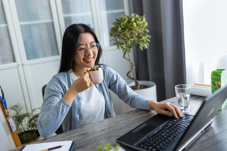 A young Thai woman enjoys a cup of coffee while participating in an online business video call from her home office.の写真素材