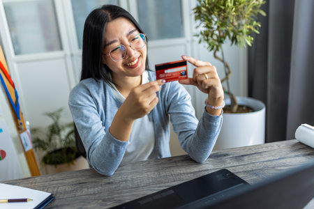 A young Thai woman is shopping online from home using her credit card while browsing on her computer.の写真素材