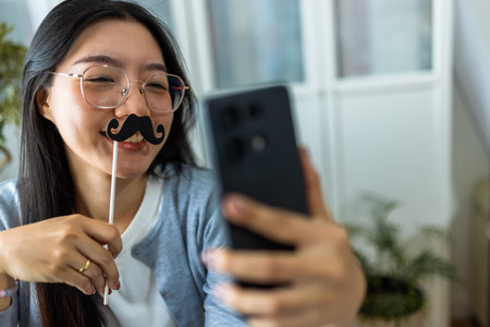 A young Thai woman sits at her desk in a home office environment, playfully wearing fake mustaches as she takes a selfie with her smartphone.の写真素材