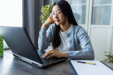 Thai woman sits at a desk in a comfortable home environment, focused on her computer as she works remotely.の写真素材