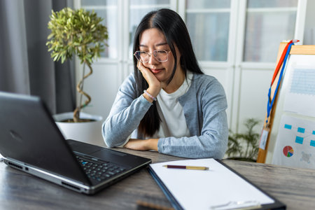 A young Thai woman sits at her desk working from home on her computer, appearing very sleepy and tired.の写真素材