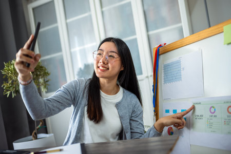 A young Thai woman works from home, delivering an online presentation using her smartphone. She actively engages her audience by pointing to a presentation displayed on a board beside her.の写真素材