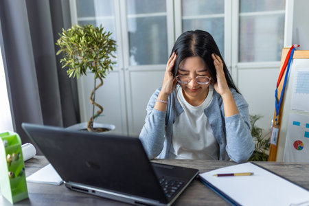Young Thai woman is working from home at her computer, visibly exhausted and holding her head in pain from a headache.の写真素材