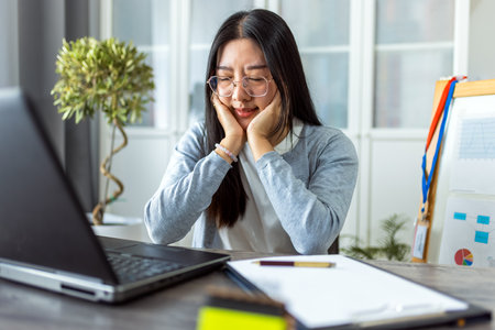 A young Thai woman sits at her desk working from home on her computer, appearing very sleepy and tired.の写真素材