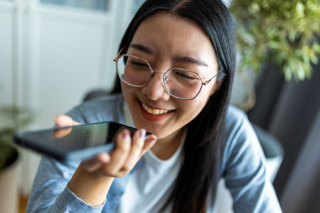 Thai woman sits at a desk in a modern home setting, multitasking as she works on her computer and sends a voice message on her smartphone.の写真素材