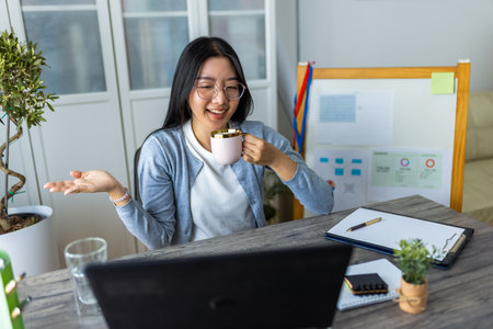 A young Thai woman enjoys a cup of coffee while participating in an online business video call from her home office.の写真素材