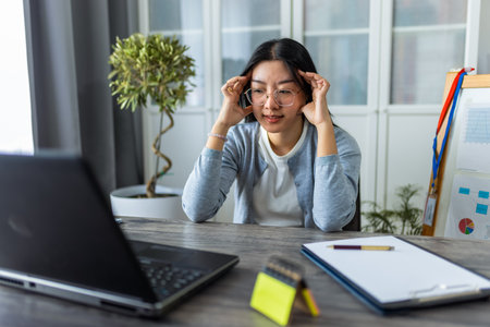 A young Thai woman is working from home at her computer, visibly exhausted and holding her head in pain from a headache.の写真素材