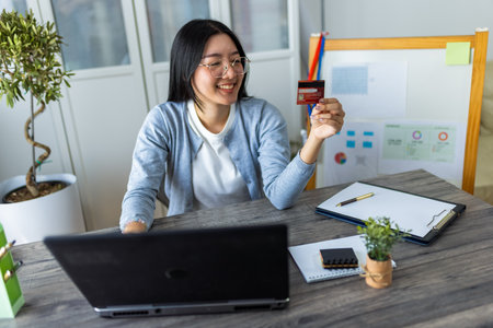 A young Thai woman is shopping online from home using her credit card while browsing on her computer.の写真素材