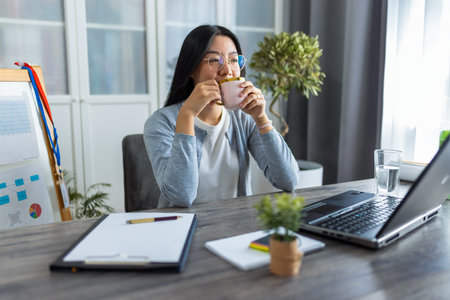 A young Thai woman enjoys a cup of coffee while participating in an online business video call from her home office.の写真素材
