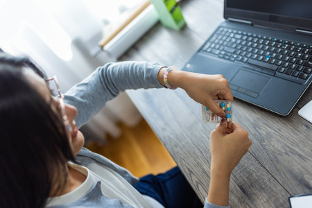 A young Thai woman works from home on her computer while taking medicine, showing a moment of self-care during her busy remote workday.の写真素材