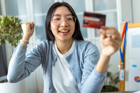 A young Thai woman is shopping online from home using her credit card while browsing on her...
